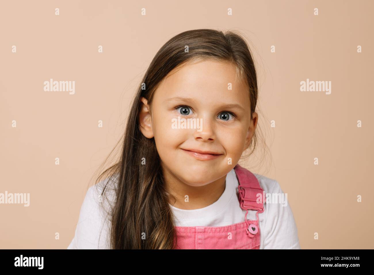 Porträt eines Kindes mit leuchtenden, leicht auskragenden Augen und einem glückseligen Lächeln, das die Kamera in einem leuchtend rosa Jumpsuit und einem weißen T-Shirt auf Beige anschaut Stockfoto