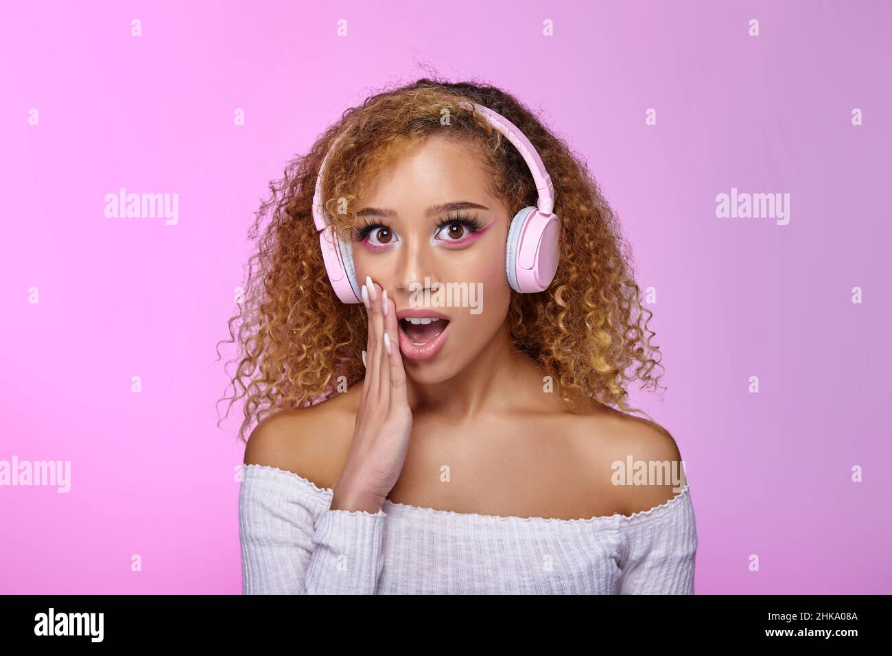 Schockierte afroamerikanische, lockige Frau, die im Studio Songs in Kopfhörern hört, während sie mit geöffnetem Mund auf einem rosa Hintergrund ihre Erstaunen ausdrückt Stockfoto
