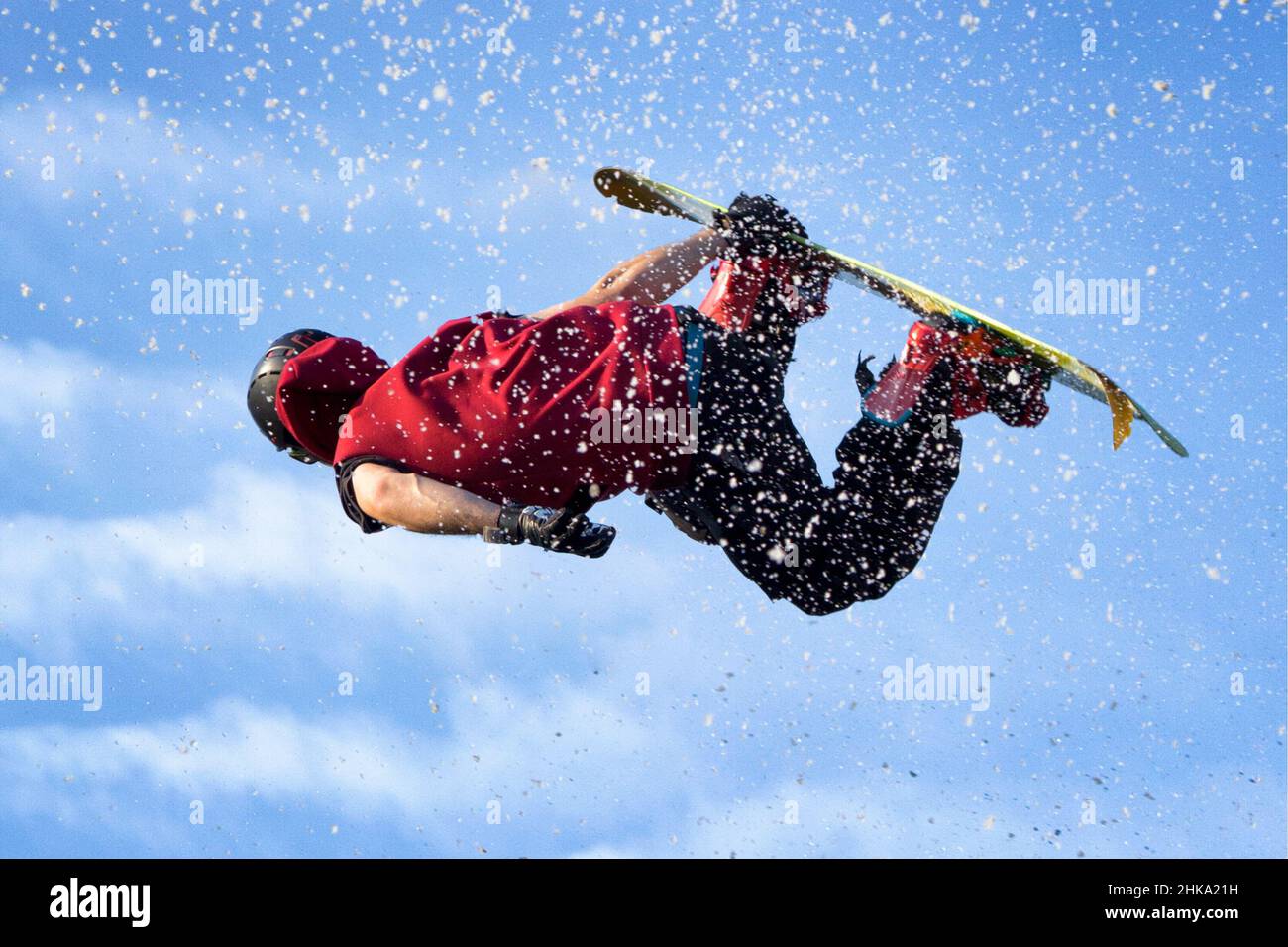 Snowboard-Jumper, der hoch in der Luft fliegt, mit schneebedecktem Staub auf blauem Himmel. Stockfoto