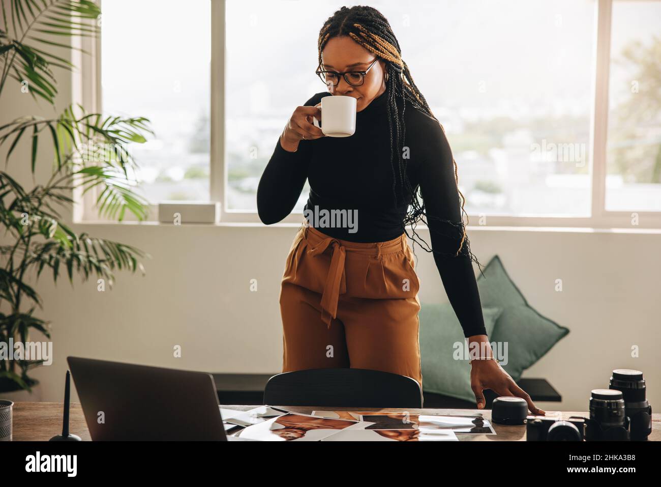 Selbstständige junge Frau, die in ihrem Heimbüro Kaffee trinkt. Kreative junge Frau, die an einem Schreibtisch mit Fotoausrüstung steht. Fotografin Stockfoto