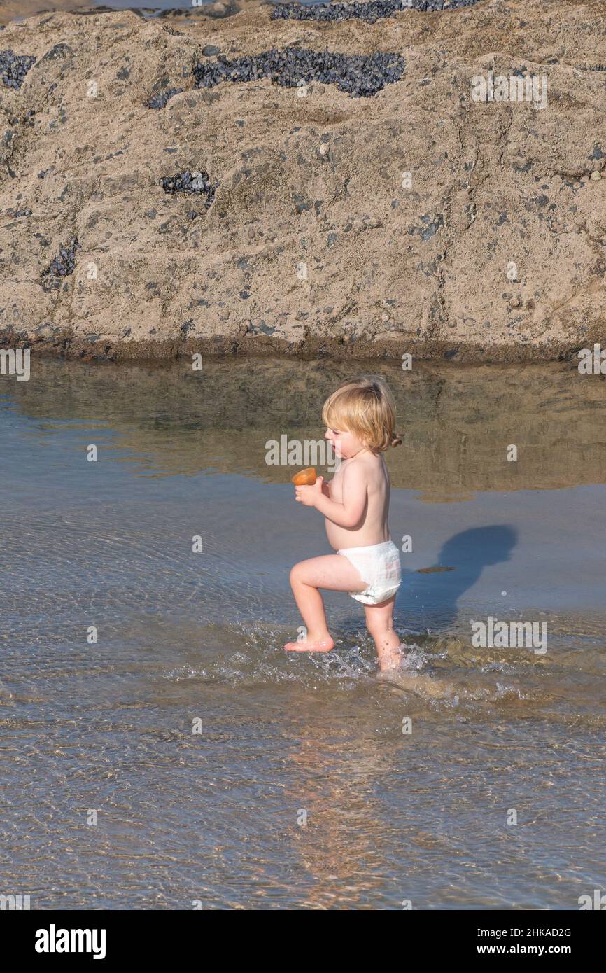 Ein sehr kleines Kind, das eine Windel trägt und Spaß beim Laufen durch ein Wasserbecken am Fistral Beach in Newquay in Cornwall hat. Stockfoto