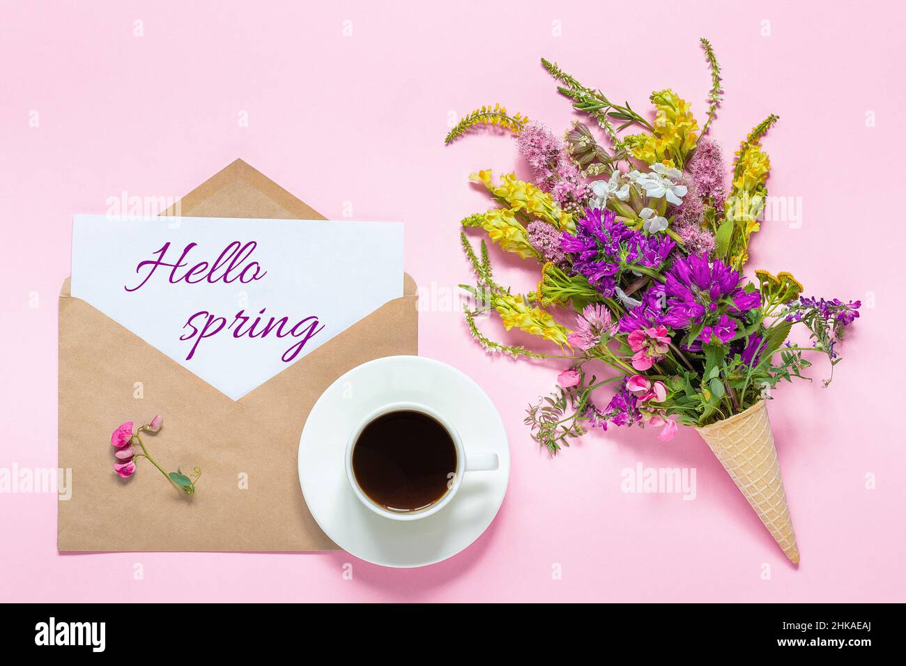 Blumenstrauß in Waffelkegel, Tasse Kaffee und Umschlag mit Kartentext Hallo Frühling auf rosa Hintergrund Grußkarte Flat Lay Draufsicht C Stockfoto