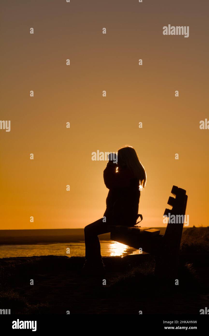 Silhouette einer jungen Frau mit Fernglas Vogelbeobachtung auf Einer Bank im Sunset Keyhaven UK Stockfoto