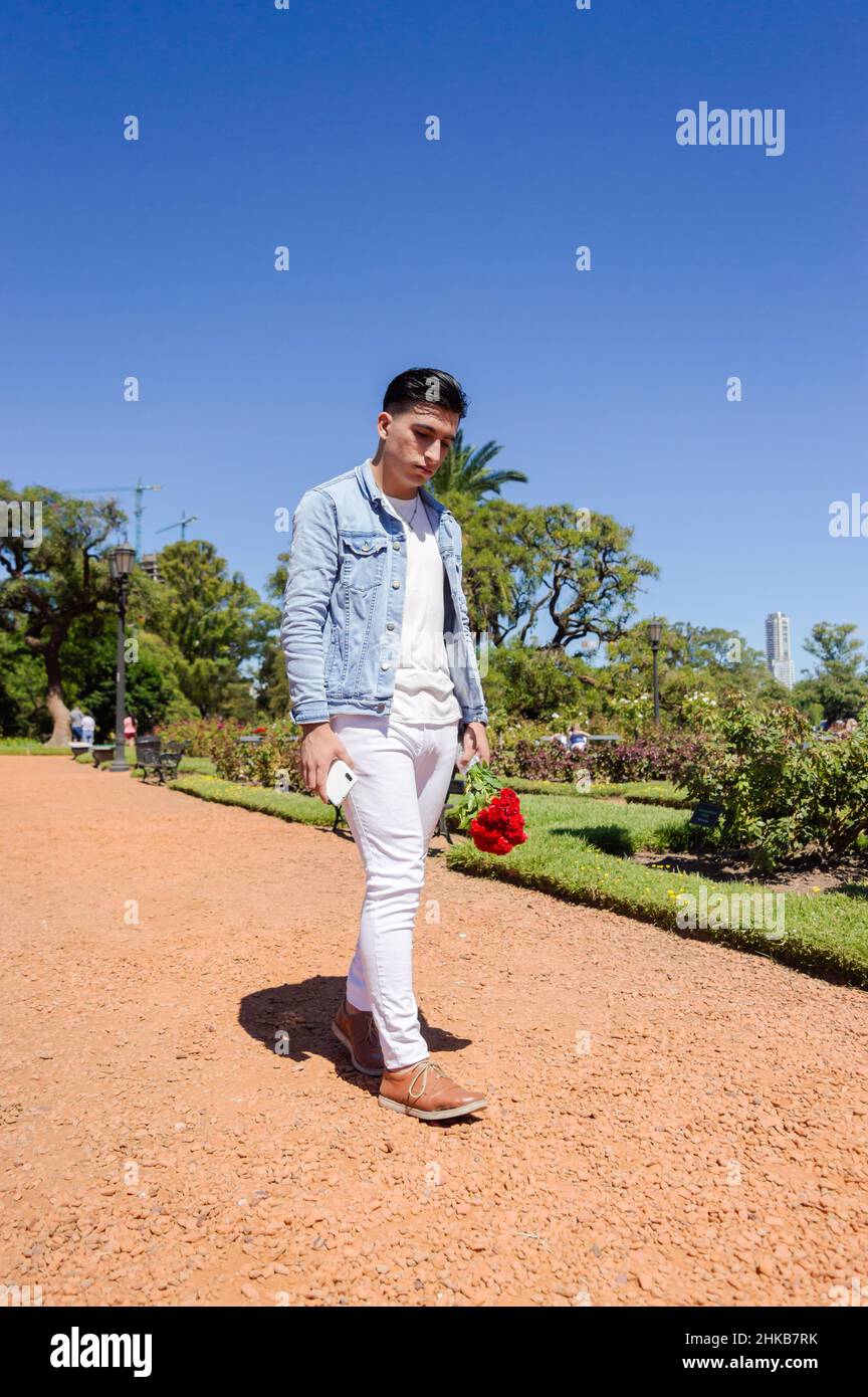 Trauriger, verhöhnter junger argentinischer Mann aus dem Kaukasus, der in einem von der Liebe besiegten Park mit einem Blumenstrauß und dem Telefon in seinem Garten nach unten geht Stockfoto