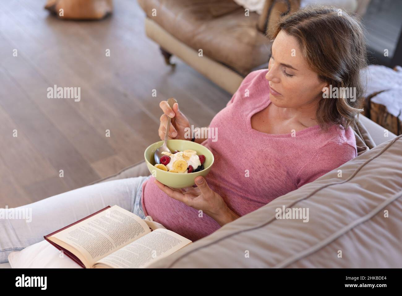 Kaukasische Schwangere, die einen Snack isst, während sie ein Buch liest, während sie zu Hause auf der Couch sitzt Stockfoto