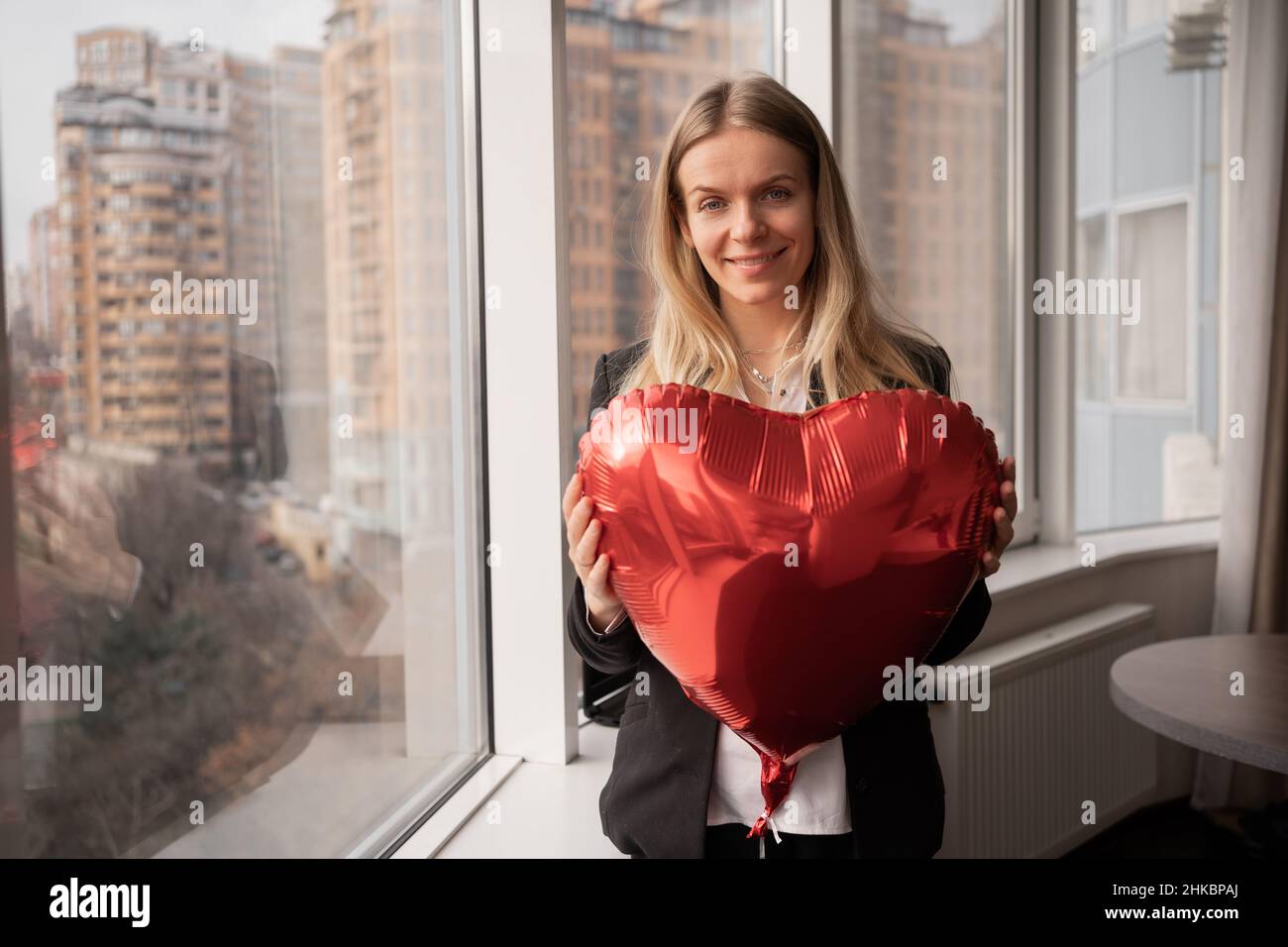 Porträt einer blonden Frau mit einem roten Ballonherz in den Händen, Büroangestellter, der den valentinstag feiert, tausendjährig am Fenster stehend und blickt Stockfoto