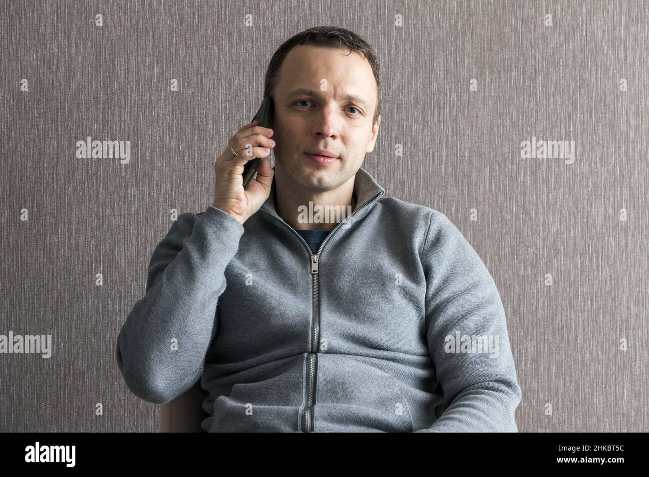 Junger erwachsener Europäer spricht auf dem Smartphone in einem Sessel in der Nähe einer leeren grauen Wand, zwangloses Studiofoto Stockfoto