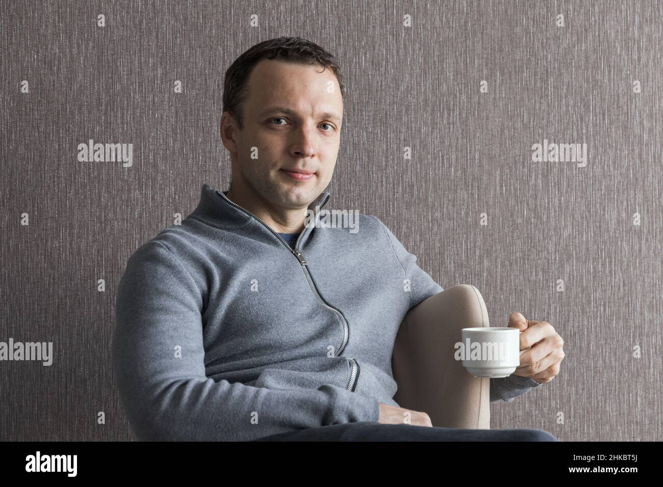 Junger Erwachsener, positiver Kaukasischer, trinkt Kaffee, sitzt in einem Sessel, legeres Studiofoto Stockfoto