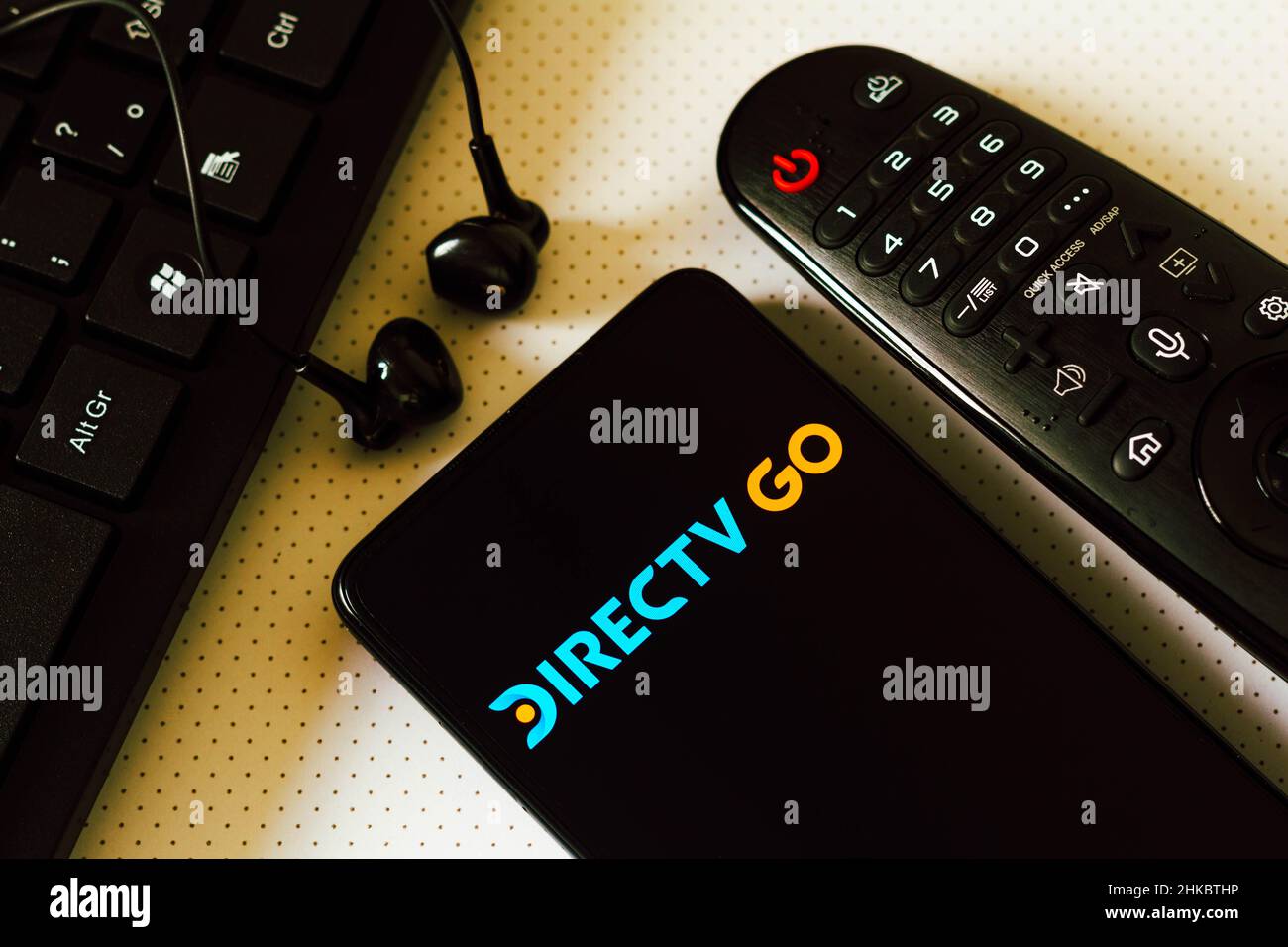In dieser Abbildung wird das Logo des DirecTV Go, ein Pay-TV-Dienst und On-Demand-Inhalte über das Internet auf einem Smartphone neben einer TV-Fernbedienung, Kopfhörern und einer Tastatur angezeigt. Stockfoto