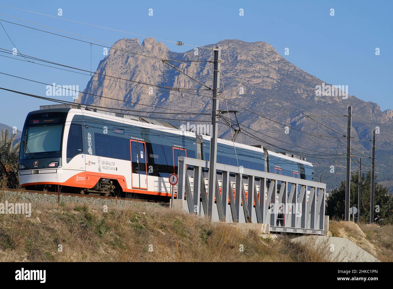 Villajoyosa Alicante Spain 01.27.22 Weißer und orangefarbener Elektrozug mit getönten Fenstern über die Stahlträgerbrücke. Stromleitungen und Masten. Berg Stockfoto
