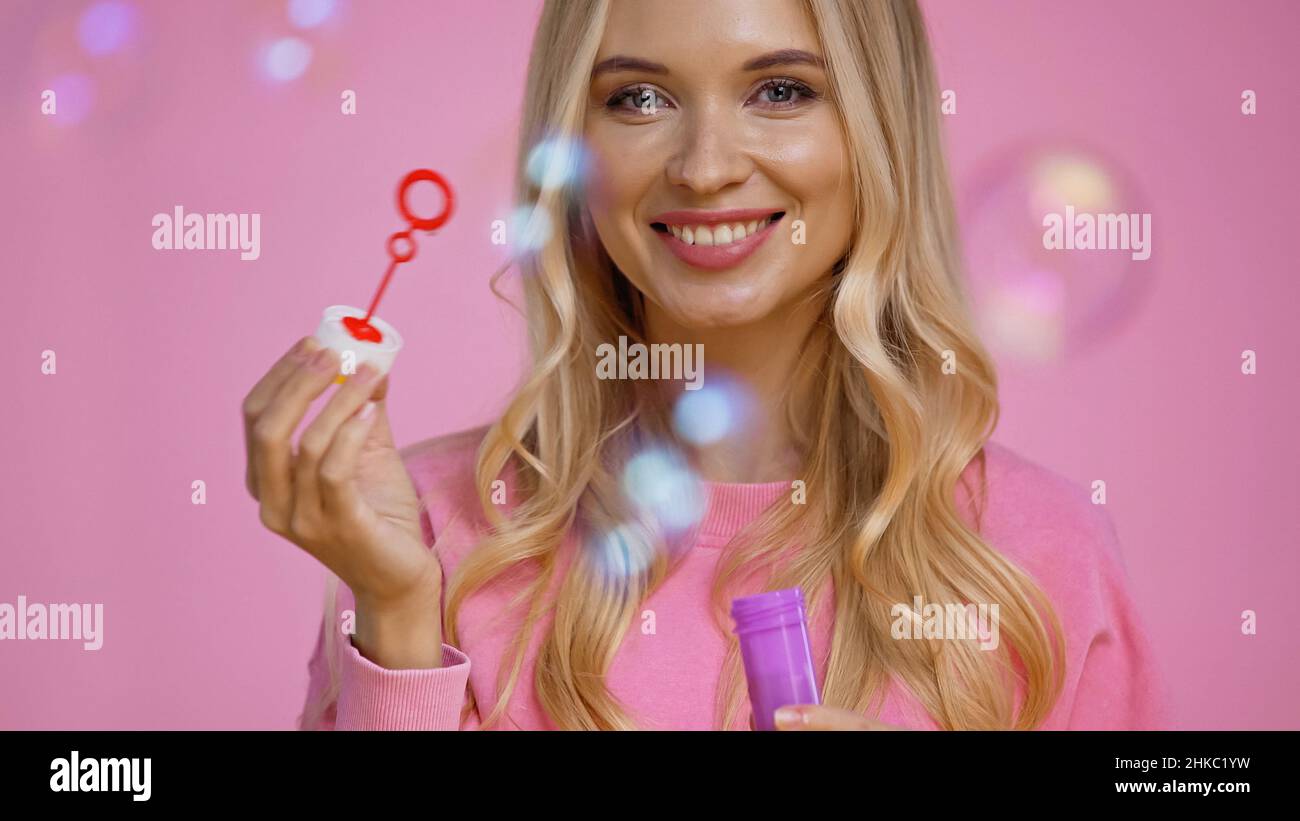 Positive blonde Frau hält Zauberstab in der Nähe Seifenblasen isoliert auf rosa Stockfoto