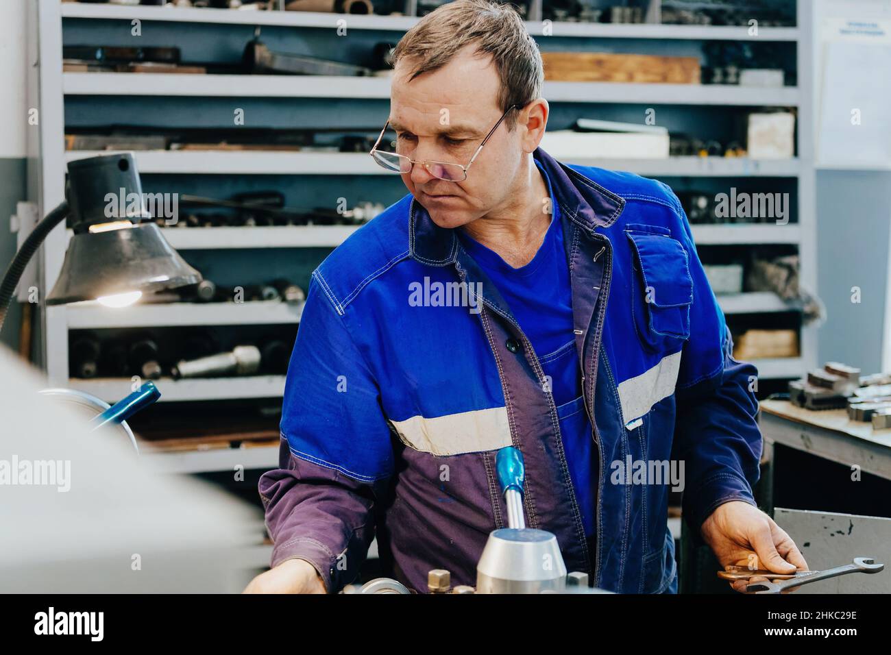 Der Erwachsene Drehmaschine von 50-55 Jahren arbeitet in der Werkstatt hinter der Drehbank. Industrielle Produktion von Metallteilen. Kaukasischer Arbeiter in Overalls. Authentische Arbeitsprozess-Szene in der Produktion. Echter Arbeiter. Nicht inszenierte Szene. Stockfoto