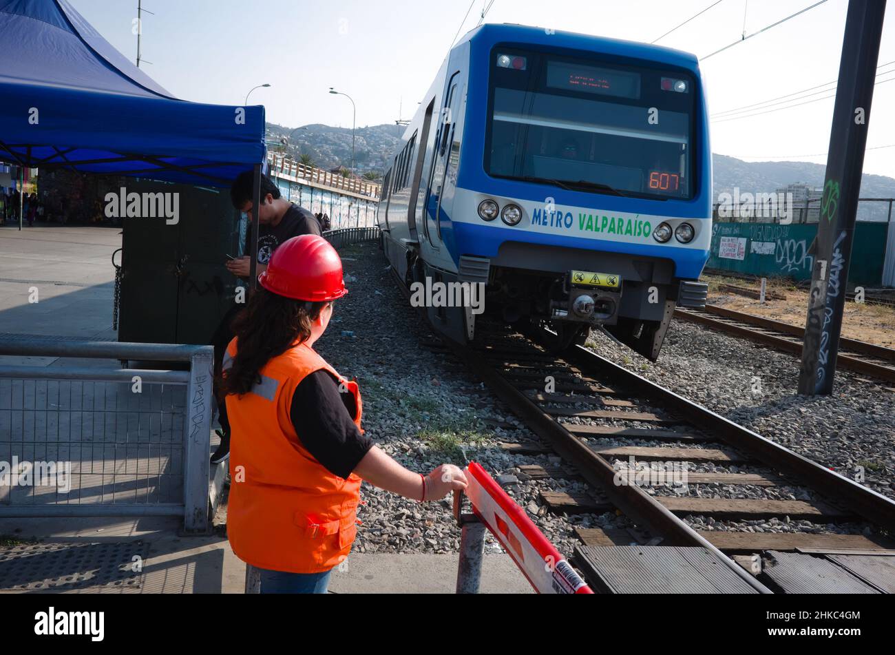 Valparaiso, Chile - Februar 2020: Frau in orangefarbener Sicherheitsweste und Schutzhelm steht an geschlossener Barriere. Die Metro-Lokomotive Valparaiso fährt vorbei Stockfoto