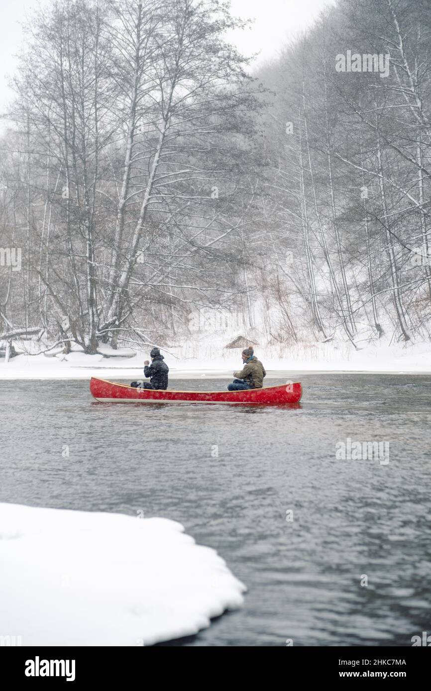 Zwei Personen paddeln in einem roten Holzkanu auf dem verschneiten Fluss. Stockfoto