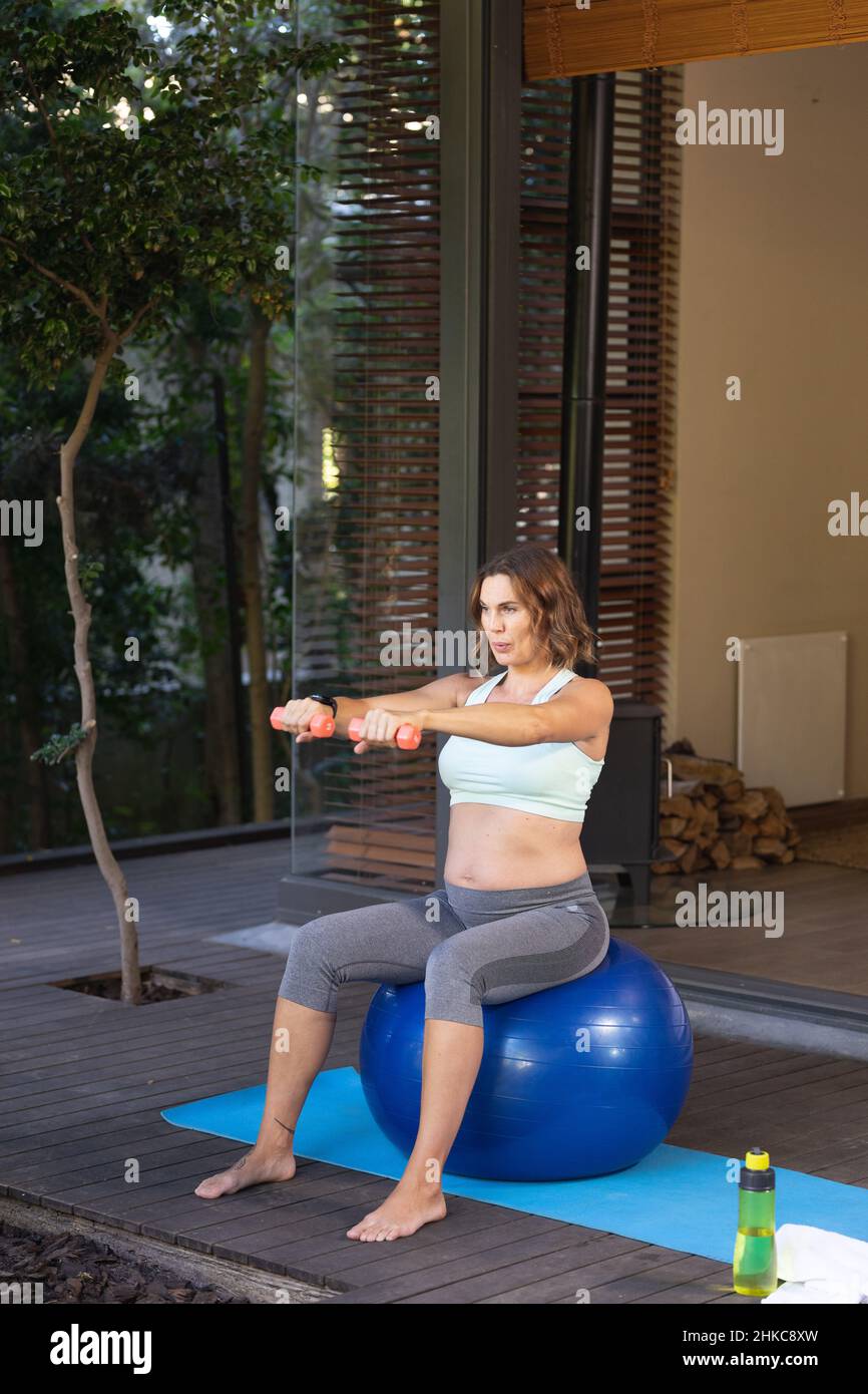 Kaukasische Schwangerin, die zu Hause mit Hanteln auf dem Übungsball trainiert Stockfoto