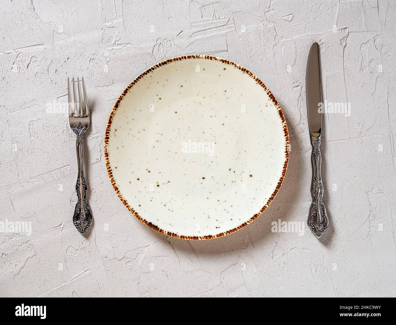 Tabelle einstellen. Weißer gesprenkelter Essteller zwischen elegantem Tischmesser und Gabel auf strukturiertem grauen Hintergrund. Leerer Porzellanteller mit braunem Rand. Stockfoto