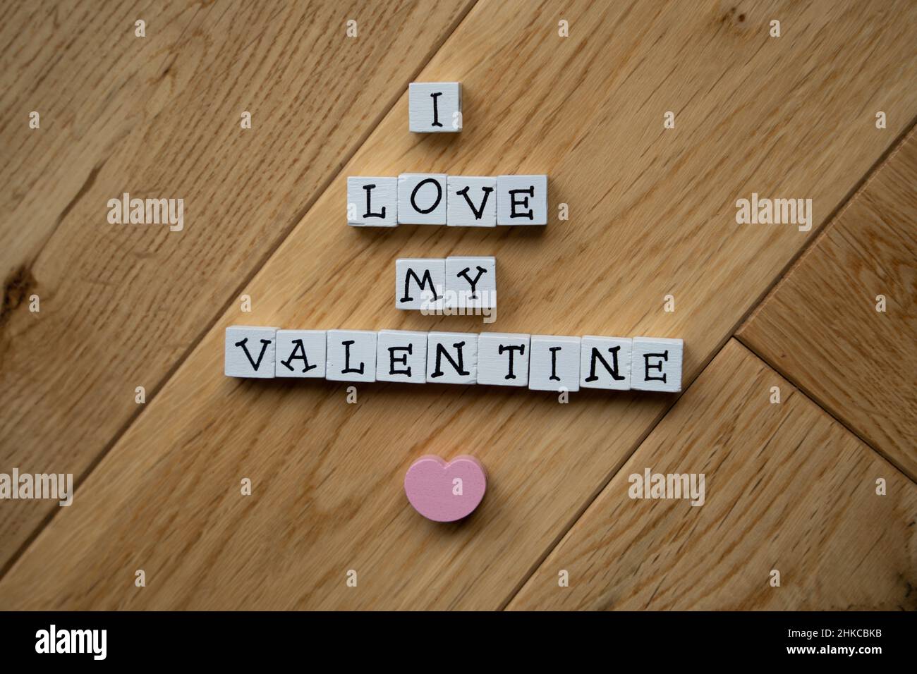 Weiße Holzbalken mit schwarzen Großbuchstaben bringen eine Botschaft zum Valentinstag mit rosafarbenem Herzen auf Holzparkett-Hintergrund zum Ausdruck. Stockfoto