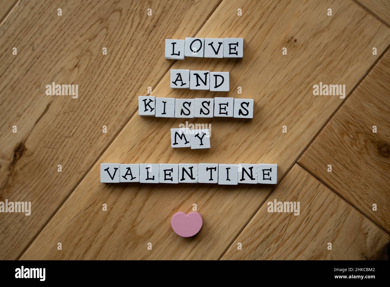 Weiße Holzbalken mit schwarzen Großbuchstaben bringen eine Botschaft zum Valentinstag mit rosafarbenem Herzen auf Holzparkett-Hintergrund zum Ausdruck. Stockfoto