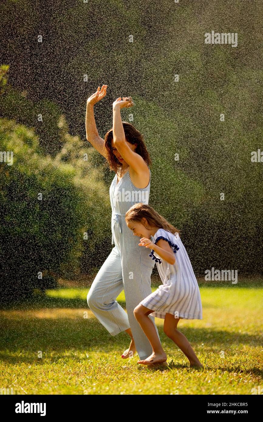 Nettes kleines Mädchen, das mit ihrer Mutter Spaß mit Wasser unter Bewässerungsregner hat Stockfoto