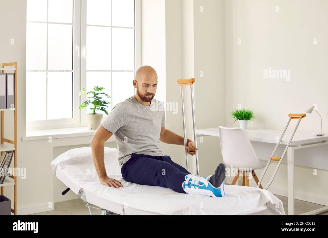 Aufgeregter junger Mann mit Knöchelverletzung sitzt während des Krankenhausaufenthalts auf der Untersuchungstisch. Stockfoto