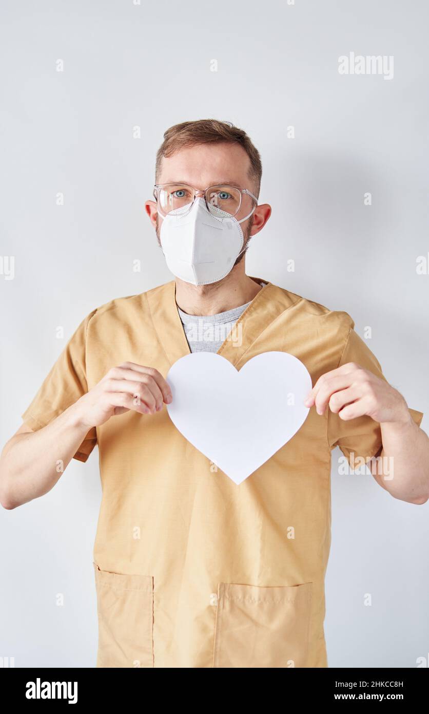 Porträt des kaukasischen männlichen Kardiologen Arzt in medizinischer Uniform und medizinische Maske mit weißem Papier Herzform. Erwachsene Arzt Chirurg im Krankenhaus posiert über der hellen Wand. Hochwertige Fotos Stockfoto
