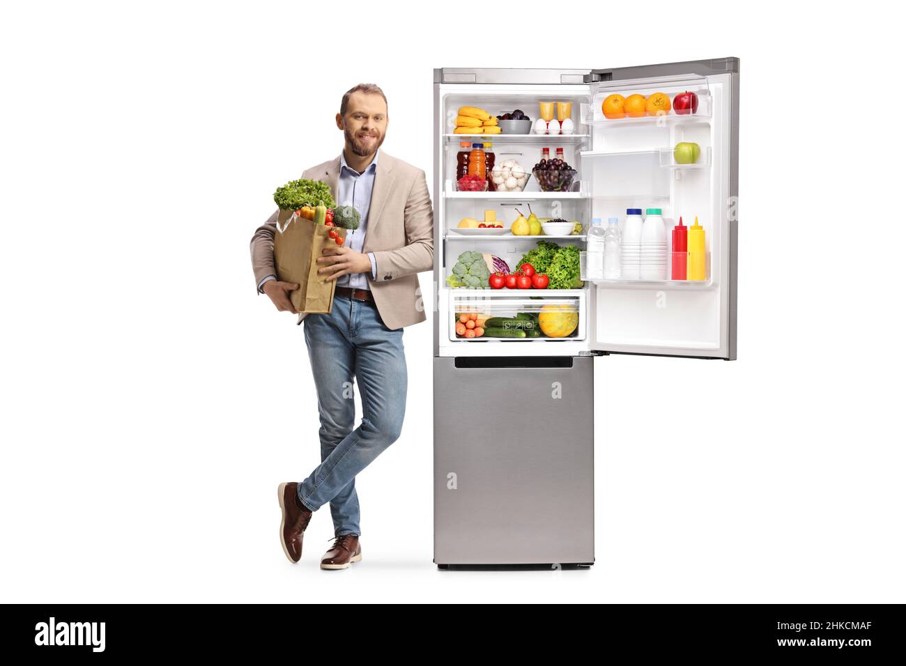 Porträt eines jungen, zwanglosen Mannes, der eine Einkaufstasche in der Hand hält und sich auf einen Kühlschrank stützt, wobei gesunde Lebensmittel auf weißem Hintergrund isoliert sind Stockfoto
