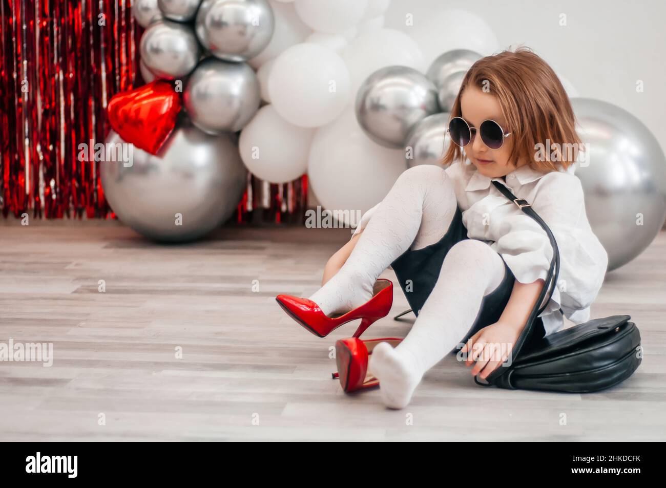 Ein modisches Baby in Sonnenbrillen versucht an den roten Schuhen ihrer Mutter. Mode und moderne Kleidung. Glitter hell metallic Spiegel Hintergrund, neue Trends V Stockfoto