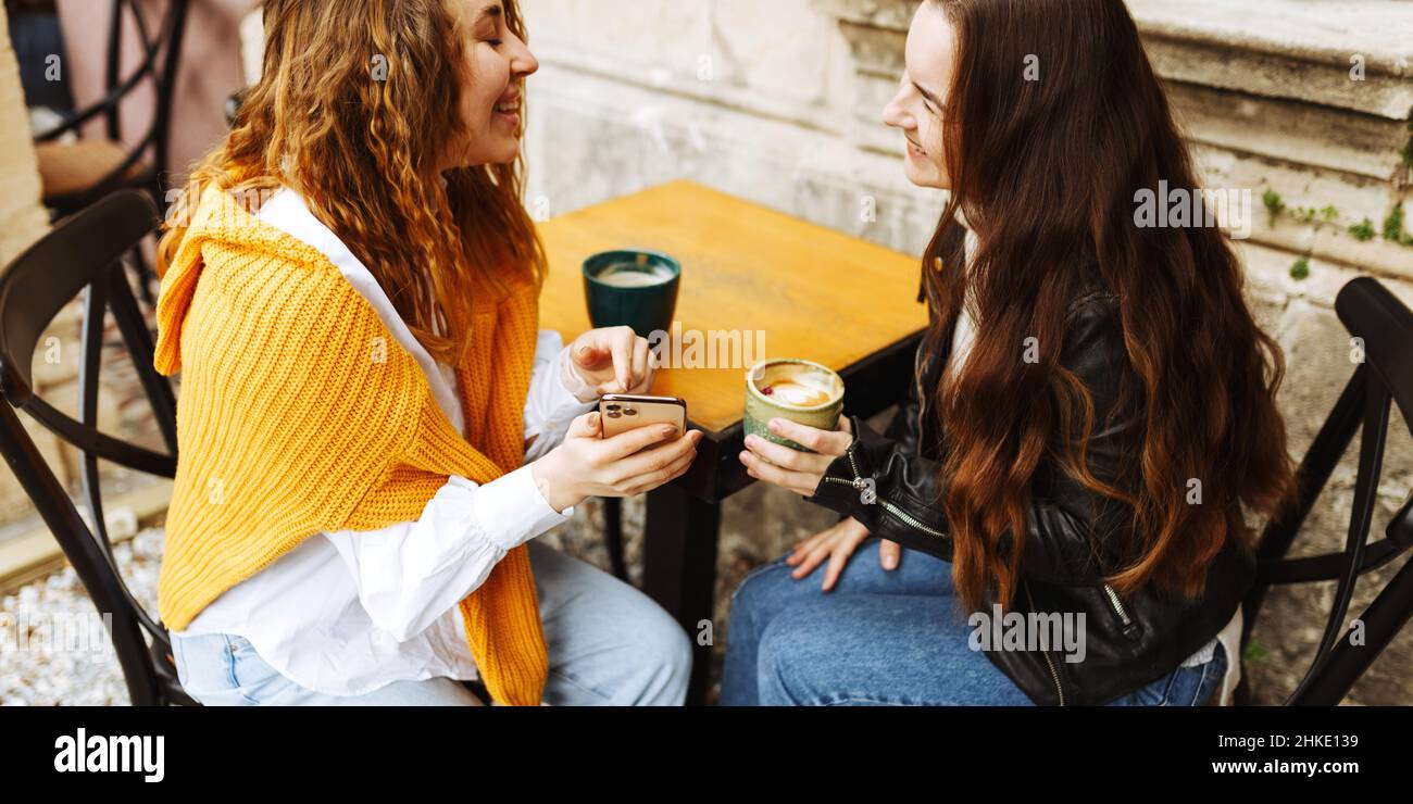 Fröhliche weibliche beste Freundin teilen Multimedia-Dateien auf Smartphones verbringen Freizeit zusammen, lächelnde Hipster Mädchen trinken Kaffee, sitzen auf der Terrasse Café. Stockfoto