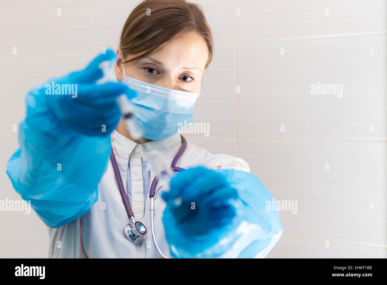 Junge Ärztin in einer medizinischen Maske und Handschuhen mit einer Spritze in der Hand und einem Stethoskop in einem Krankenhaus während einer Coronavirus-Pandemie. Selektive Stockfoto
