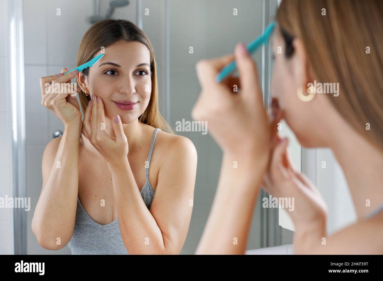 Entfernung von Gesichtshaaren. Schöne junge Frau, die ihr Gesicht mit einem Rasiermesser zu Hause rasiert. Hübsche Frau mit Rasiermesser im Badezimmer. Stockfoto