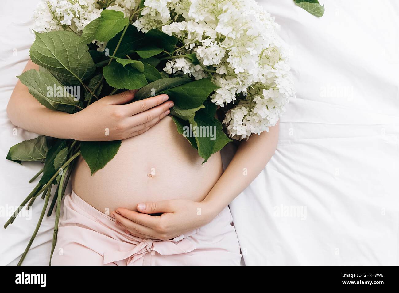 Nahaufnahme von nicht erkennbaren Händen einer Schwangeren, die sanft den Bauch berühren und auf einem bequemen Bett mit schönen frischen Sommerblumen liegen. Zukünftige Mutter Stockfoto