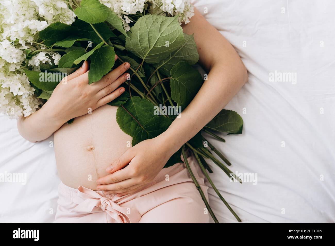 Cropped shot schöne junge schwanger weiblich 20s liegend auf bequemen weißen Bett mit frischen Frühlingsblumen, genießt wunderbare Morgen und Schwangerschaft. Kopieren Stockfoto