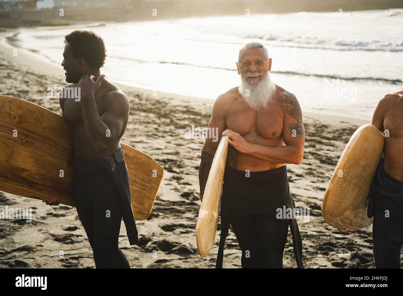 Surfer-Freunde mit mehreren Generationen, die nach der Surfstunde am Strand Spaß haben – konzentrieren Sie sich auf das Gesicht eines älteren Mannes Stockfoto