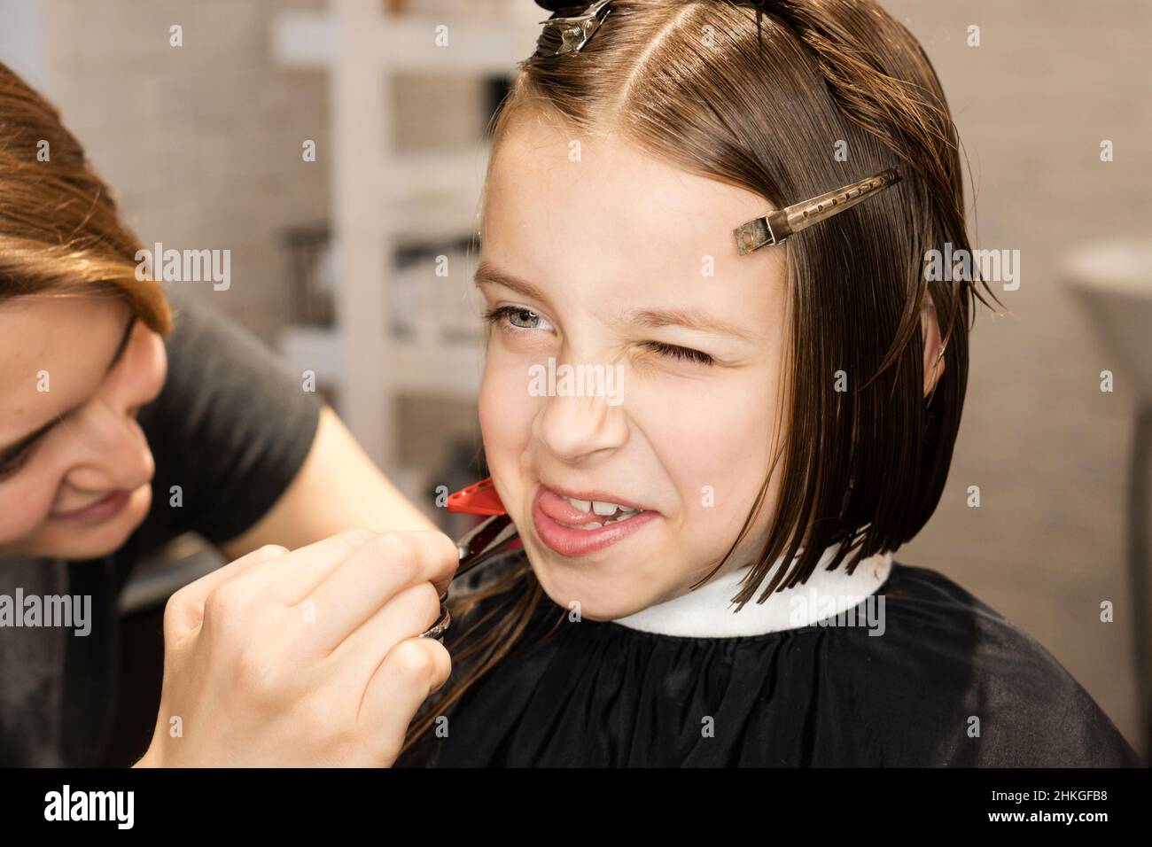 Wunderschöne glückliche kurzhaarige kleine Mädchen macht Grimassen in Friseurladen in Prozess des Schneidens der Haare in Schönheitssalon. Frisur für Kinder. Moderne Kinder Kurz Bob Haarschnitt Stockfoto