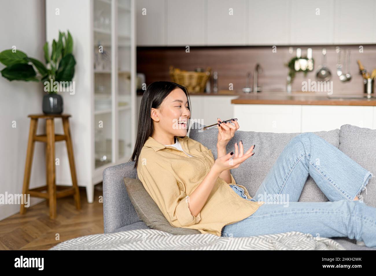 Charmante junge Frau, die zu Hause auf dem Sofa sitzt, Sprachnachrichten aufnimmt und sendet. Frau, die die Spracherkennungs-App auf dem Mobiltelefon nutzt und im Mikrofon des Smartphones spricht Stockfoto