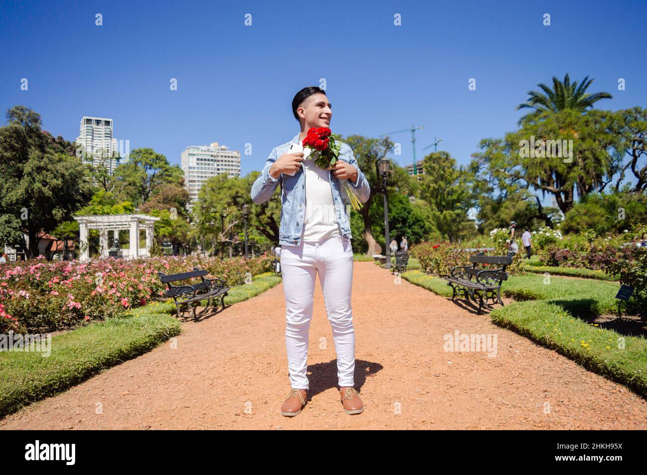 Der junge kaukasische hispanische Latino-Mann in einem Stadtpark mit einem schönen Garten fixiert seine Jacke, mit einem Blumenstrauß in der Hand wird er glücklich gehen Stockfoto