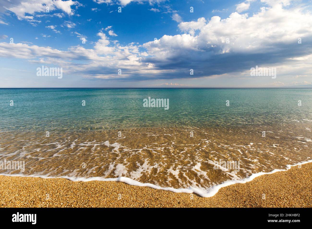 Ruhiges türkisfarbenes Meer und Wolken am blauen Himmel in Italien, Europa. Kleine Wellen an einem Sandstrand im Vordergrund. Stockfoto