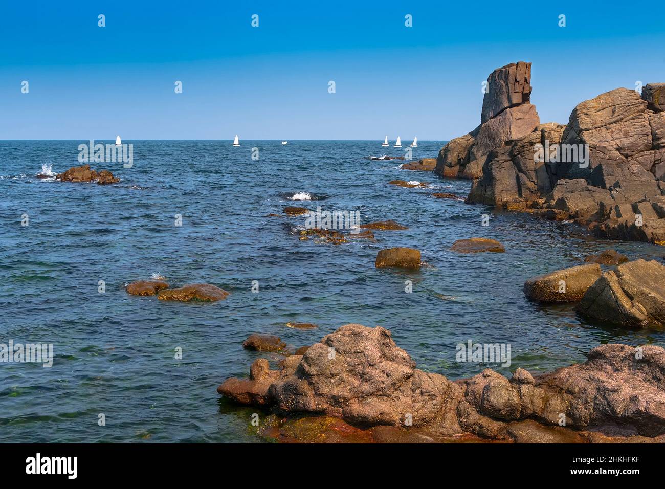 Felsige Küste mit Steinblöcken, die aus dem Wasser ragen, und Segelboote am Horizont Stockfoto