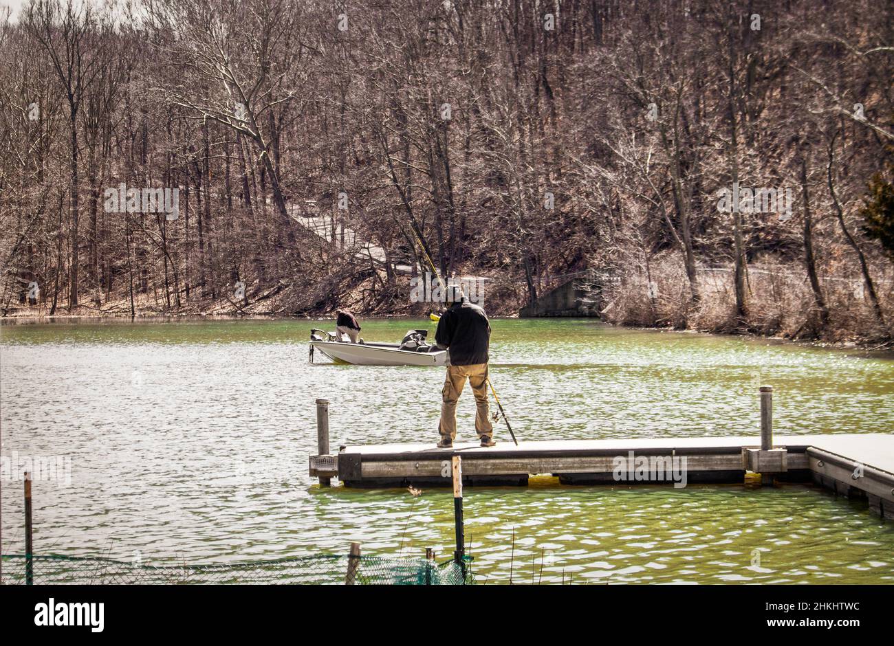 Mann steht auf dem Dock Angeln, während ein anderer Mann in einem Bass-Boot schwimmt vorbei auf einem kleinen See im frühen Frühjahr Stockfoto