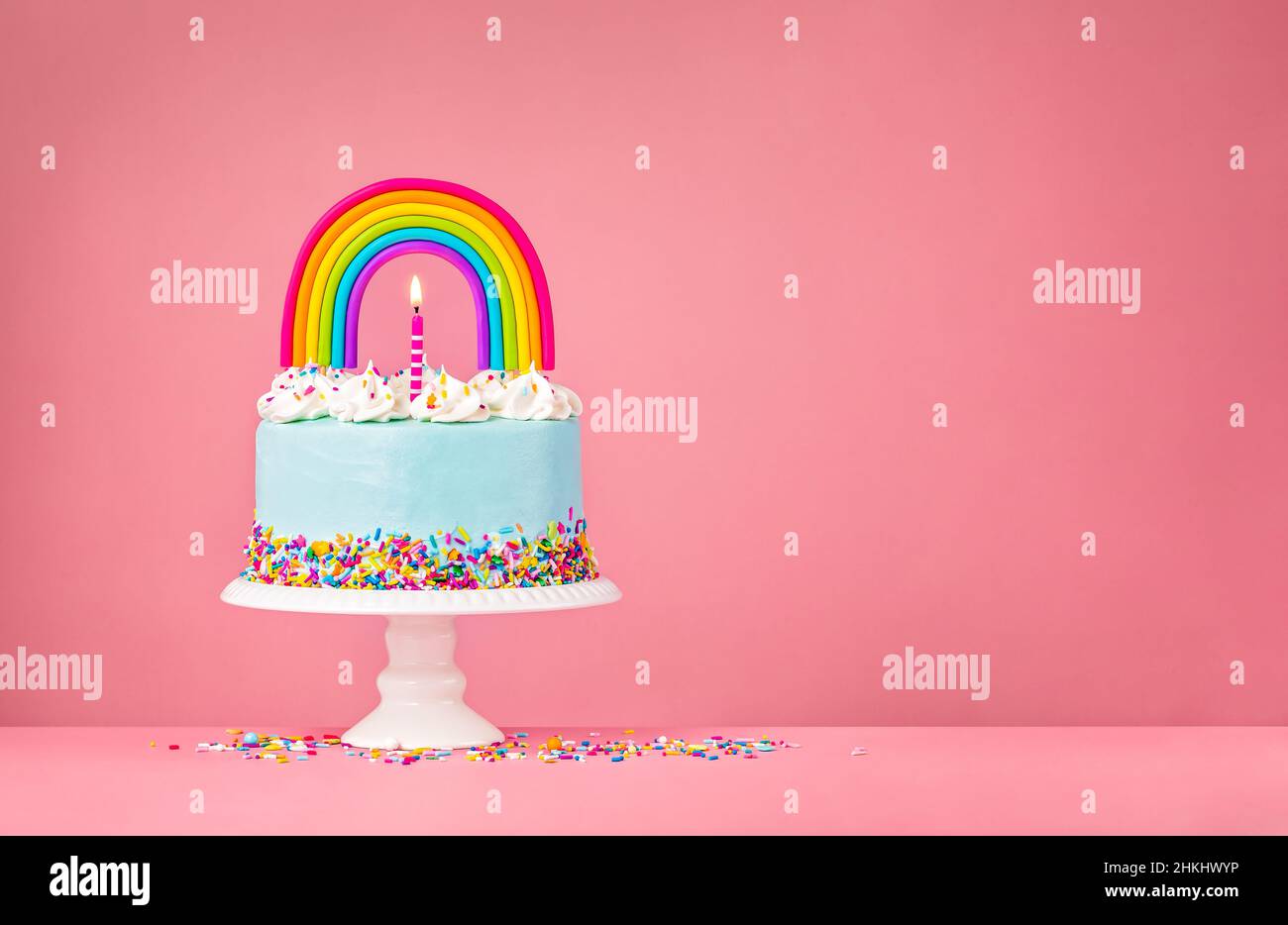 Blue Birthday Cake mit brennenden Kerzen, Streuseln und einem farbenfrohen, hausgemachten Regenbogentoper auf einem rosa Hintergrund. Stockfoto