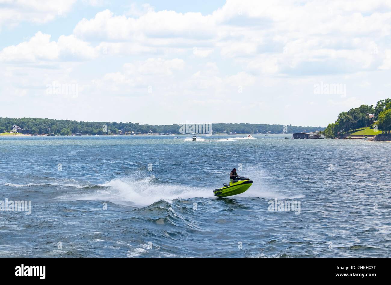 Person auf einem grünen PWC fängt Luft auf See mit anderen Booten und PWCs und Häusern und dockt am Ufer im Hintergrund Stockfoto