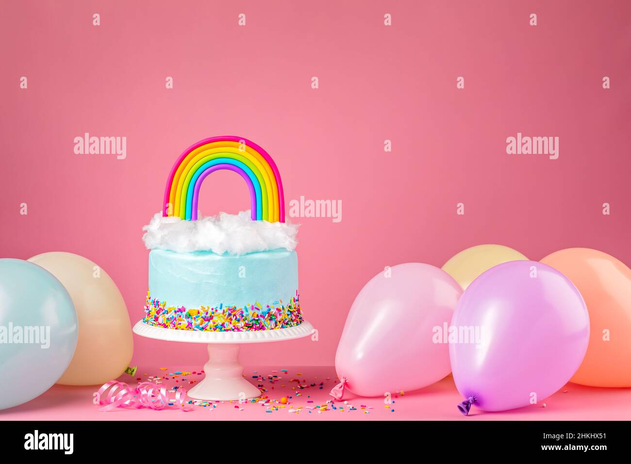 Blauer Geburtstagstorte mit Zuckerwolken aus Baumwolle und einem bunten, hausgemachten Regenbogentopf auf einem rosa Hintergrund, umgeben von Party-Ballons und Konfetti sp Stockfoto