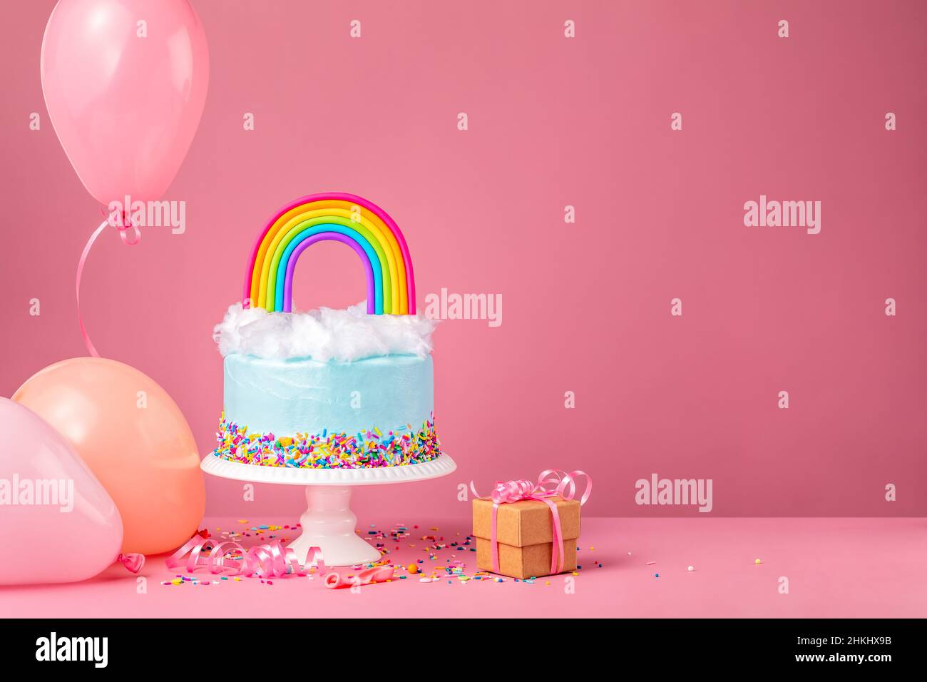 Blauer Geburtstagstorte mit Zuckerwatte und einem bunten Regenbogen auf einem rosa Hintergrund, umgeben von Partyballons, Konfetti-Streuseln und einem Geschenk. Stockfoto