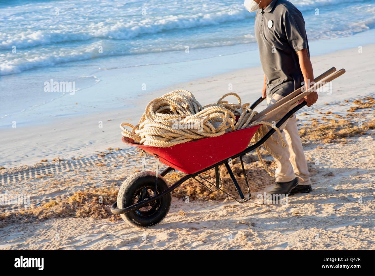 Ein Latino mit einer covid19 Maske schiebt eine Schubkarre mit einem Seil an einen tropischen Strand in Mexiko Stockfoto