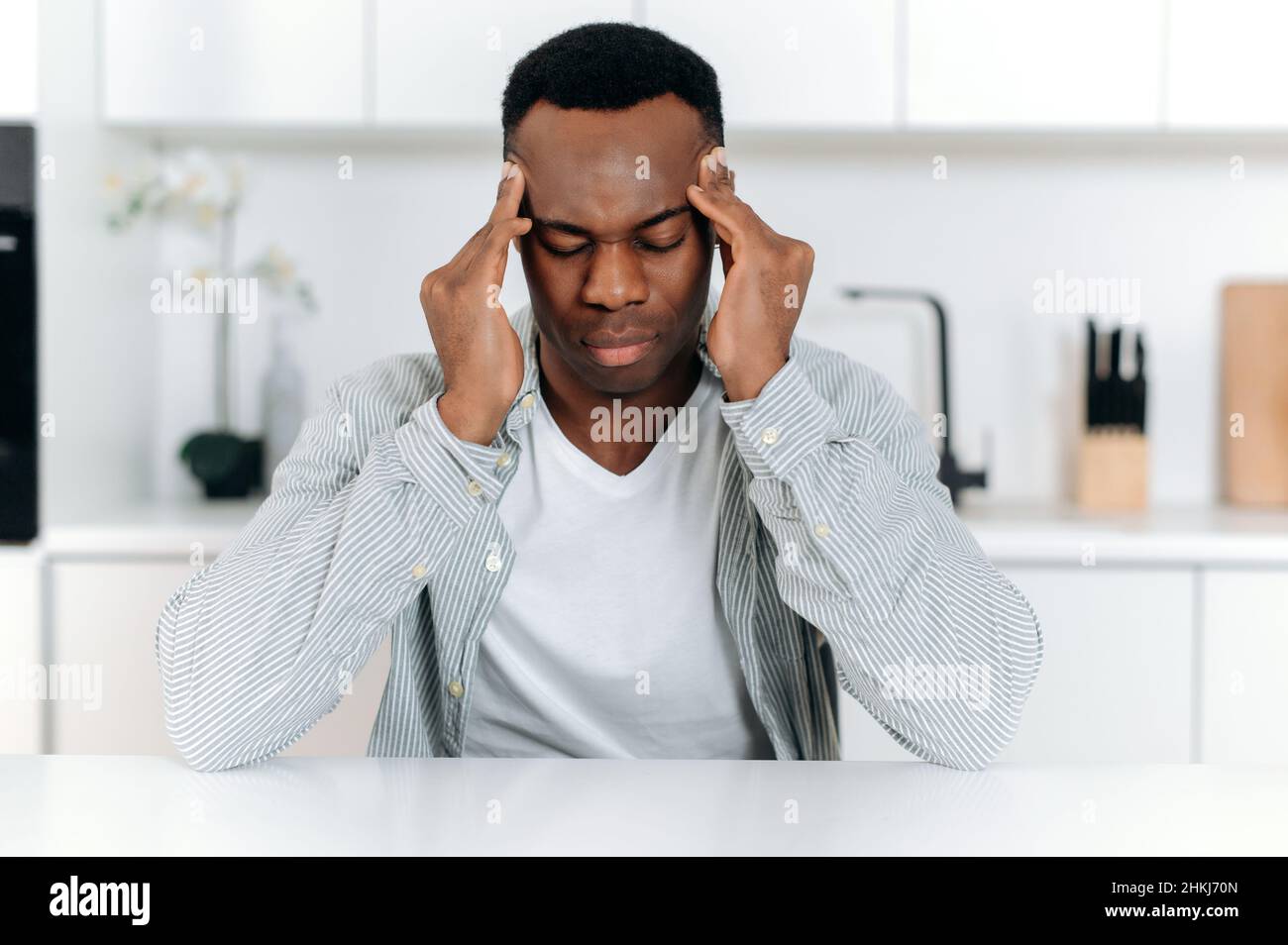 Trauriger afroamerikanischer Typ in Stress, allein am Tisch sitzend, in lässiger Kleidung, Müdigkeit, Kopfschmerzen, Migräne, Massierte seine Schläfen, schließt seine Augen, braucht Ruhe, Medikamente und Behandlung Stockfoto