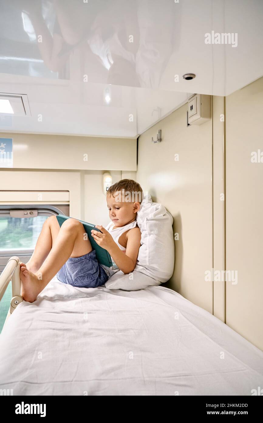 Der blonde kleine Junge sieht sich am Morgen Cartoons über einen Tablet-PC an und lehnt sich auf ein weiches Kissen im oberen Regal im Zugabteil Stockfoto