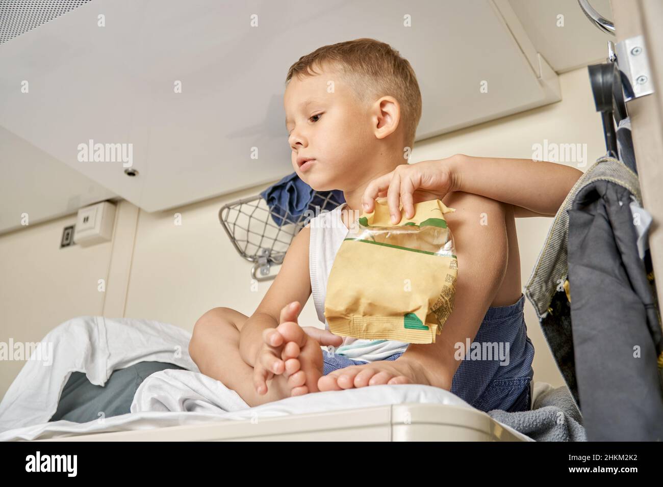 Der blonde kleine Junge mit Weste und Shorts isst Snacks aus der Papierpackung, die auf dem obersten Regal des Wagens liegt, der mit der Familie unterwegs ist Stockfoto