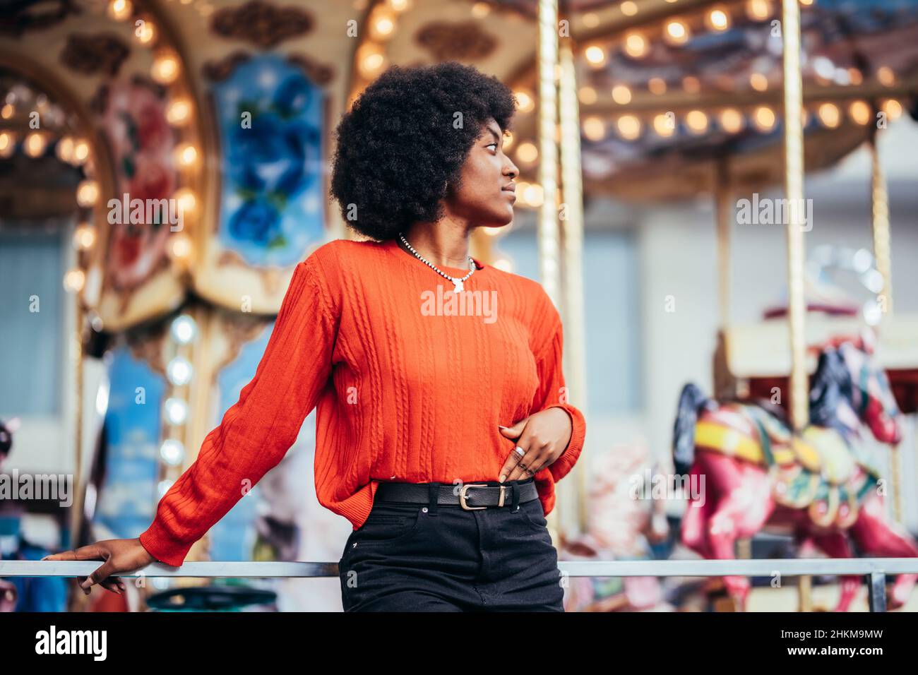 Nachdenkliche junge schwarze Frau mit afro-Frisur und rotem Pullover neben einem Karussell auf der Straße Stockfoto
