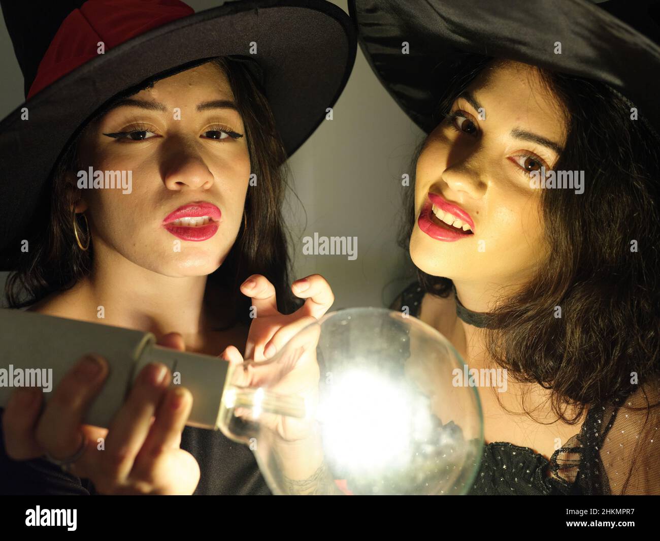 Zwei Latina-Freunde, die an halloween als Hexen gekleidet mit einer Glühbirne in ihrer Nähe auf die Kamera schauen und sie betrachten. Stockfoto