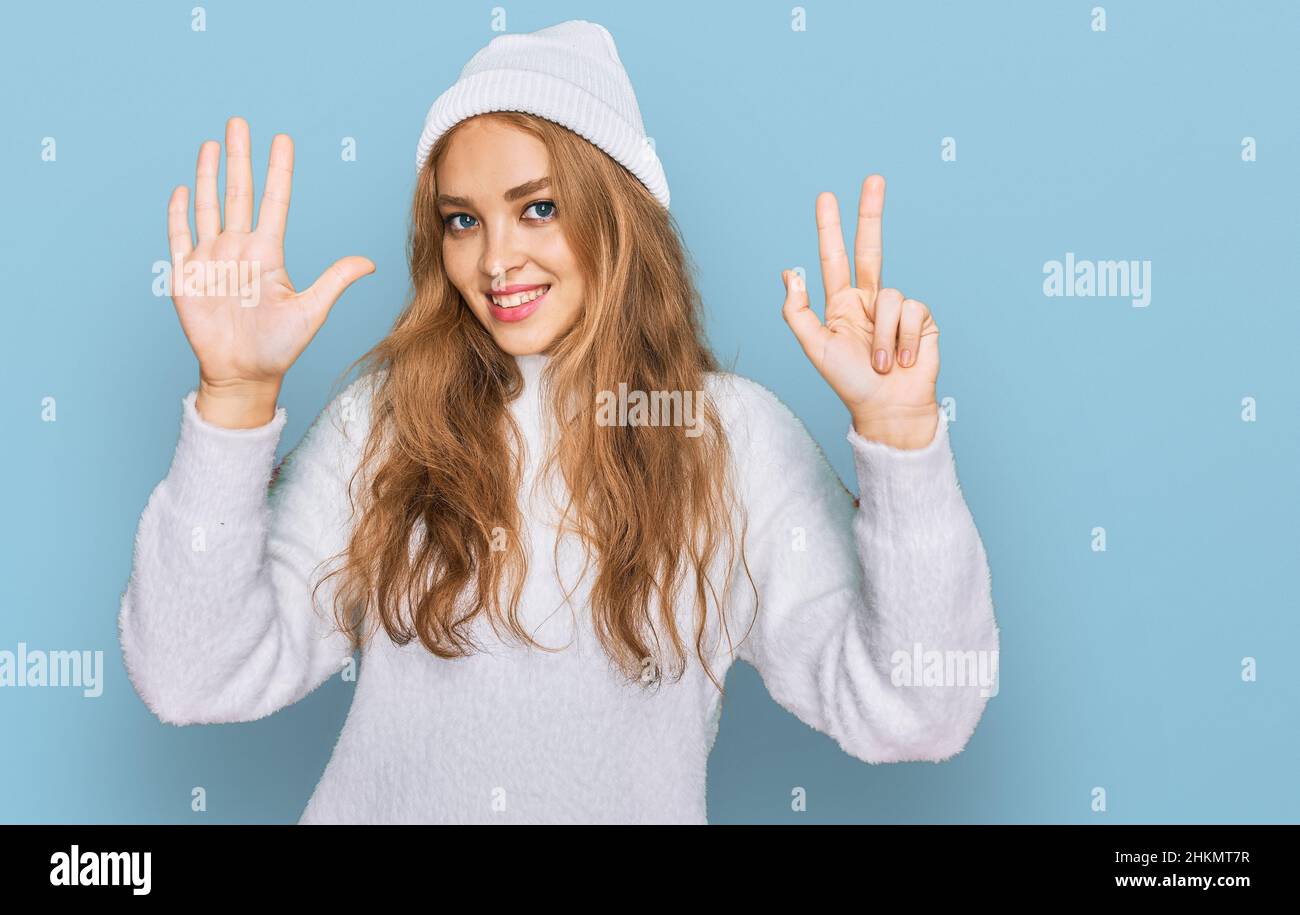 Das junge kaukasische Mädchen trägt einen Wollpullover und eine Wintermütze und zeigt mit den Fingern Nummer acht nach oben, während es selbstbewusst und glücklich lächelt. Stockfoto